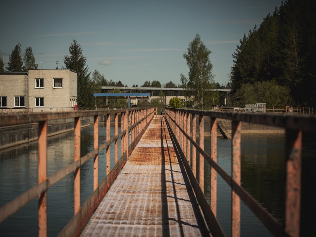 A rusty pedestrian bridge stretches over water in an industrial area with trees in Lithuania.